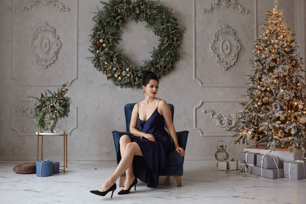 Adorable woman in a stylish dark-blue dress in front of Christmas tree. Lovable female model in trendy dress posing in the interior decorated for New year holidays - Фото, изображение