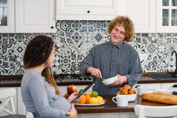 Joyful pregnant woman with dreadlocks drinks tea and watching her curly husband cooking breakfast for her in kitchen - Photo, image