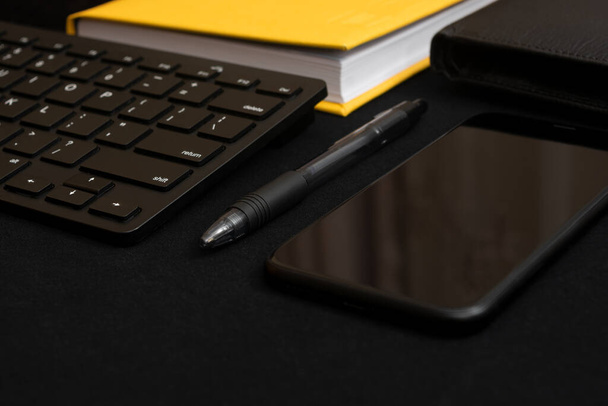 Composition with black pen, computer keyboard, smartphone, leather wallet and bright yellow notebook on dark black surface. Business concept. Business people items. Free space for text. Low key photo. Stationery. Items of successful people. Black and - Foto, Imagen