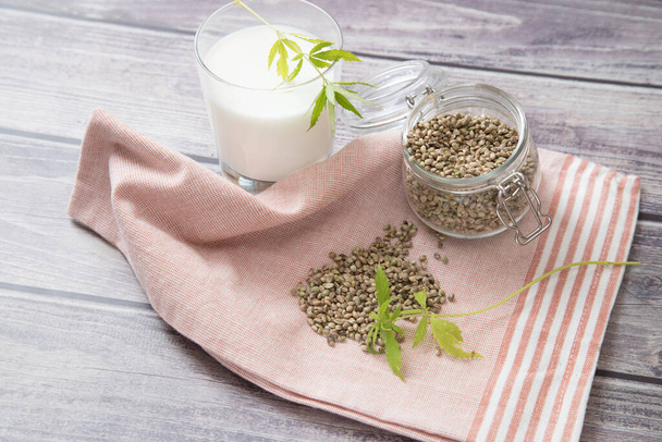 Hemp seeds on a napkin and an open glass jar with hemp milk in a glass on the table. Vegan product - Фото, изображение