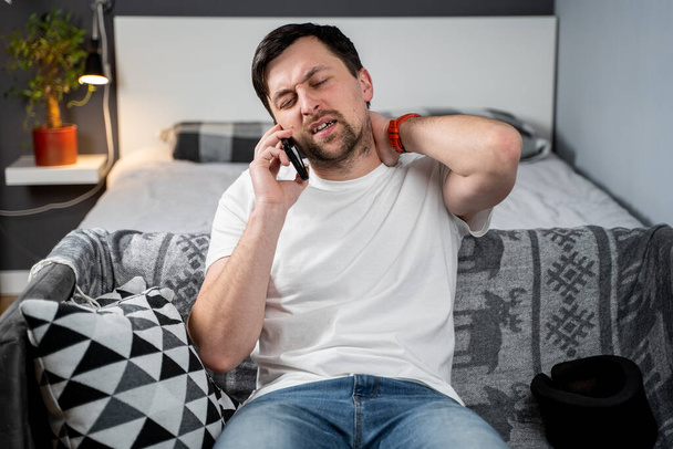 Disabled man with neck brace talking on phone with doctor, experiencing pain while sitting on couch in the living room at home. Injured young man using cervical brace makes calls using mobile phone. - Photo, Image