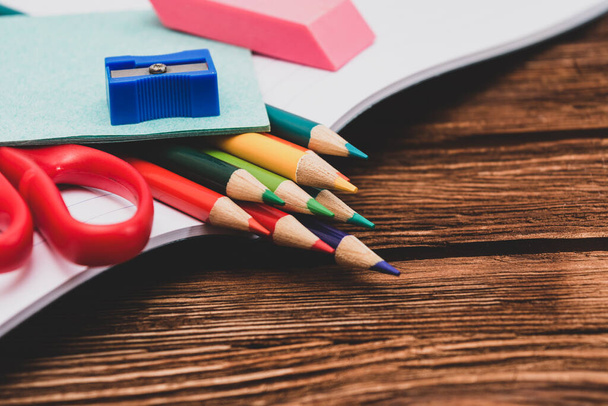 Colored pencils, sharpener, scissors and sheets for notes in an open notebook on a dark brown wooden surface. Flat lay. Back to school. Education. Office tool. Empty place for text. - Foto, Imagem