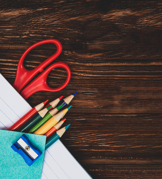 Colored pencils, sharpener, scissors and sheets for notes in an open notebook on a dark brown wooden surface. Flat lay. Back to school. Education. Office tool. Empty place for text. - Photo, Image