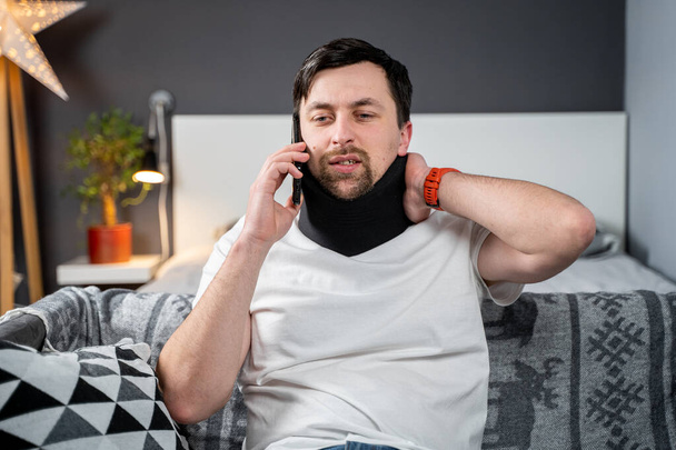 Disabled painful man with neck brace talking on phone at home. Man with spine trauma in neck brace cervical call insurance company for medical help. Male with neck injury calls doctor after accident. - Photo, Image