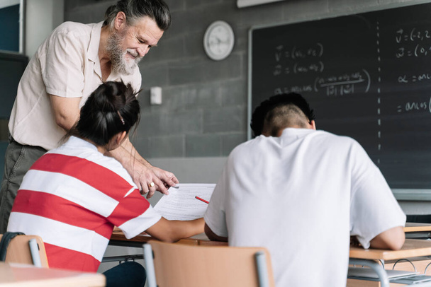 Mature Professor explaining maths exercise to teenager students. Friendly Teacher with beard helping student in secondary school classroom - Fotografie, Obrázek