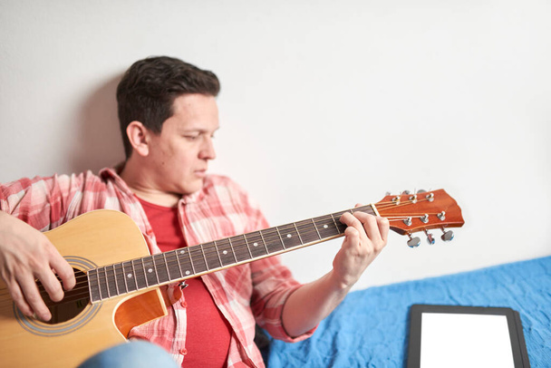 Młody człowiek uczy się grać na gitarze w domu za pomocą tabletu. Koncepcje e-learningu, technologii, hobby i muzyki. - Zdjęcie, obraz