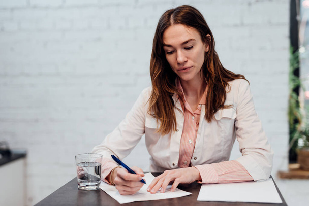Γυναίκα γιατρός κάθεται στο γραφείο της και γράφει με ένα στυλό σε ένα φύλλο χαρτί - Φωτογραφία, εικόνα