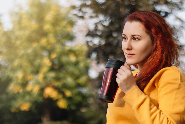 Young red-haired woman drinks coffee from reusable thermo mug. Enjoy autumn sunny day in nature. Eco-friendly lifestyle. - Photo, Image