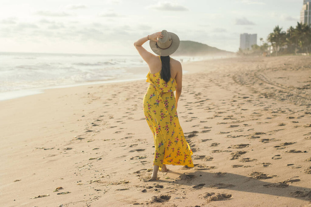 Mujer joven adulta caminando en la playa, mano izquierda levantada, mano derecha en sombrero - Foto, imagen