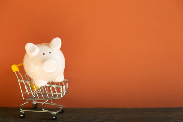 Pink piggy bank in shopping cart on wooden desk. red wall background - Photo, Image