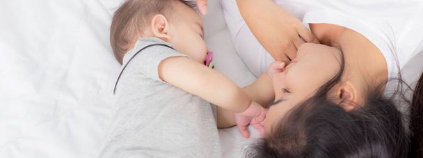 Young asian mother touch head of little baby girl with tender on bed in the bedroom, mom love newborn and care, woman with expression with child together, parent and daughter, family concept. - Fotó, kép