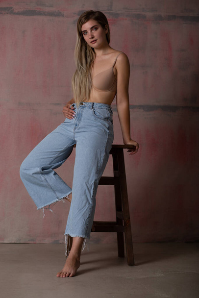 Sexy fashionable blonde posing in a bra and jeans. A girl with a perfect slender body. - Фото, изображение