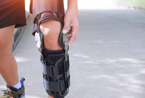 Close up of young man's hand turning angle movement control of supportive knee brace on his leg while jogging in public park - Photo, Image