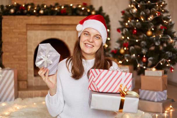 Good looking happy optimistic female wearing white sweater and red santa hat holding many present boxes in hands, celebrating new year eve, posing near fireplace and xmas tree. - Φωτογραφία, εικόνα