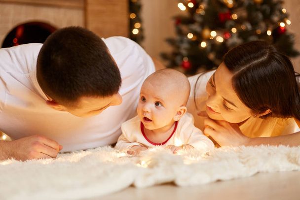 Счастливая пара с ребенком лежит на полу на белом мягком ковре, родители смотрят на своего ребенка с большой любовью, позируя в помещении с елкой на заднем плане, С Новым годом. - Фото, изображение