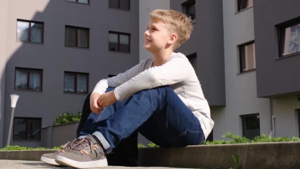 Iskolásfiú ül a járdaszegélyen jó hangulatban, és beszél valakivel a napsütéses napon. - Felvétel, videó
