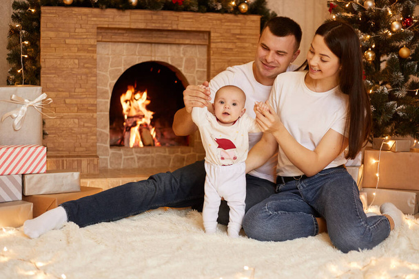 Smiling mother and father spending time together with their infant daughter while sitting on floor near fireplace and X-mas tree, merry Christmas and happy new year. - 写真・画像
