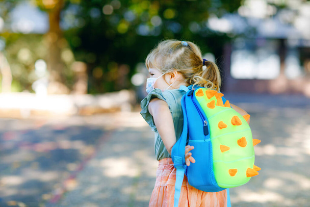 Маленькая девочка-малышка в свой первый день на пути в детскую школу в медицинской маске против коронного ковидного вируса. Здоровый красивый ребенок идет в детский сад и детский сад. Счастливый ребенок с рюкзаком - Фото, изображение