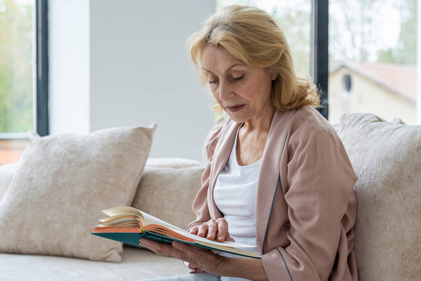 Elderly woman resting at living room. She is reading novel  - Φωτογραφία, εικόνα
