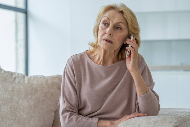 Une femme âgée triste parle sur un téléphone portable à la maison assis sur le canapé - Photo, image