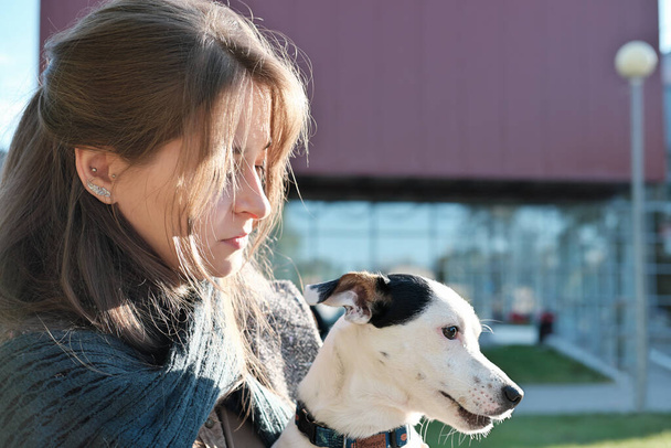 mujer joven con abrigo abrazando a su perro mientras está sentada en un banco en una ciudad. Jack Russel Terrier de rodillas de mujer mirando a un lado. cuidado de mascotas, caminar juntos y descansar al aire libre. - Foto, imagen