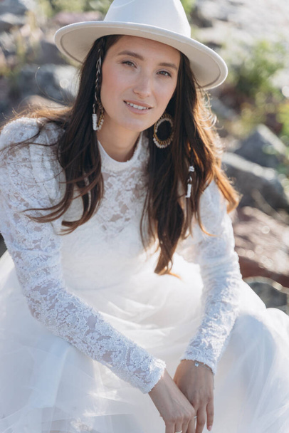 The portrait of beautiful woman dressed in a delicate white dress and with a white hat on river shore near woods - Photo, Image