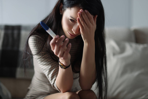 Upset woman holding pregnancy test while sitting on couch - Foto, imagen