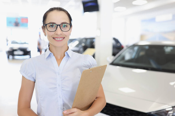 Młoda uśmiechnięta kobieta menadżer w okularach trzyma schowek na tle dealera samochodowego zbliżenie - Zdjęcie, obraz
