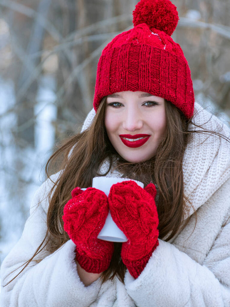 Portrait de belle fille souriante en bonnet tricoté rouge et mitaines avec tasse dans les mains. En plein air. Cadre vertical. - Photo, image