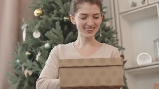 Tilting up of young beautiful Caucasian woman sitting with legs crossed on floor in foreground of decorated tree, opening box with Christmas gift, then getting excited and smiling - Footage, Video
