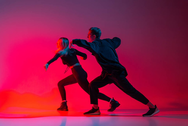 Side view horizontal image of young couple dancing hip-hop in stylish black clothes isolated over gradient pink purple background in neon light - Photo, Image