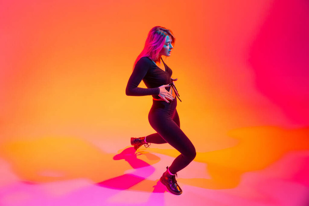 Full-length portrait of beautiful sportive Caucasian girl dancing hip-hop isolated over gradient pink yellow background in neon light - Foto, Imagem