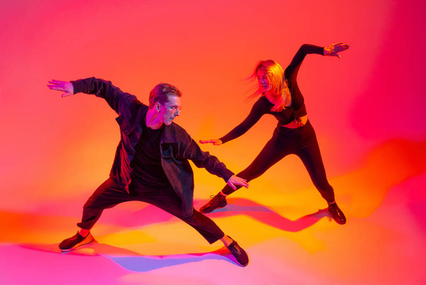 Porträt von zwei jungen männlichen und weiblichen Hip-Hop-Tänzern in Bewegung isoliert über dem Farbverlauf rosa gelb Hintergrund in Neonlicht - Foto, Bild