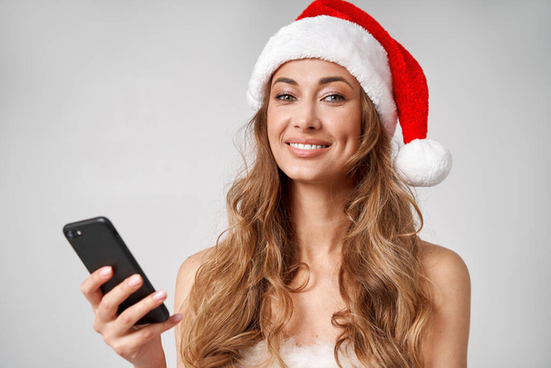 Woman christmas Santa Hat white studio background with smartphone in hand Beautiful caucasian female curly hair portrait. Happy person positive emotion Holiday concept with copy space Teeth smiling  - Foto, afbeelding