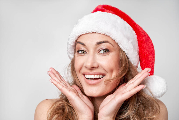 Woman christmas Santa Hat white studio background  Beautiful caucasian female curly hair portrait Happy person positive emotion Holiday concept Close up face portrait with hand - Zdjęcie, obraz