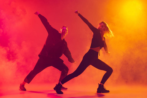 Vista inferior de dois jovens dançarinos de hip-hop em movimento isolados sobre gradiente de fundo amarelo-rosa em luz de néon em fumaça - Foto, Imagem