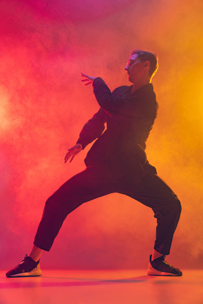 Young Caucasian man in stylish black clothes dancing hip-hop isolated over gradient pink yellow background in neon light in smoke - Foto, afbeelding