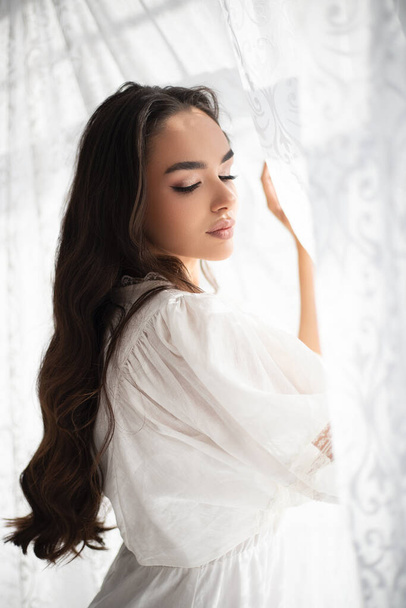 Young pretty brunette girl with incredible makeup in white summer dress among white tenderness tulle standing near the window. Indoor photography. - Zdjęcie, obraz