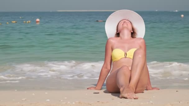 Портрет молодой улыбающейся женщины, сидящей на пляже в жёлтом купальнике и белой шляпе - Кадры, видео