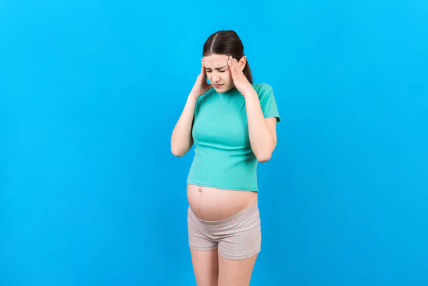Mooie jonge zwangere vrouw voelt hoofdpijn op gekleurde achtergrond geïsoleerd. Gestresste zwangere vrouw of moeder problemen. - Foto, afbeelding