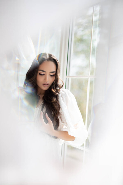 Reflection in the mirror of young pretty brunette girl with incredible makeup in white summer dress. Indoor photography. Blurred photography. - Photo, Image