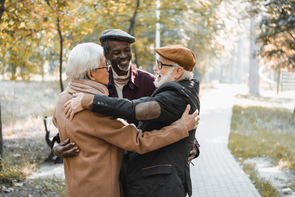 alegre anciano hombre abrazando asiático y africano americano amigos en otoño parque  - Foto, imagen