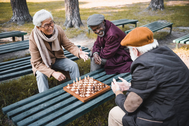 Cheerful interracial men looking at friend near chess in park  - Фото, изображение