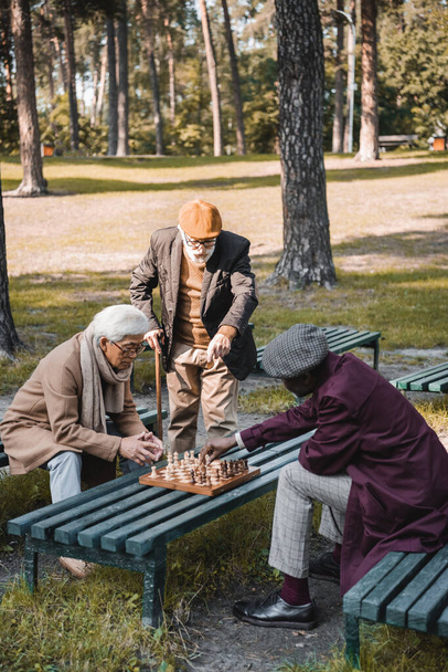 Anciano señalando con el dedo al tablero de ajedrez cerca de amigos interracial en el parque de otoño  - Foto, Imagen