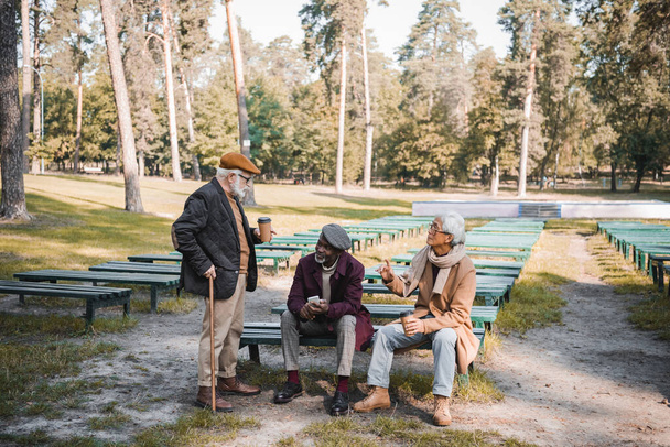 Multiethnic men with smartphone and coffee to go talking in autumn park  - Photo, Image