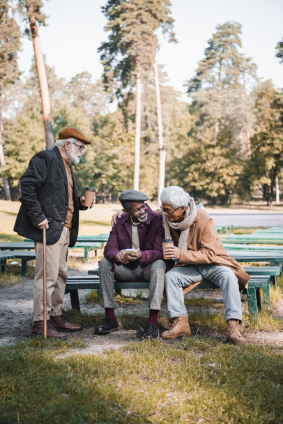 Веселые мультиэтнические мужчины с бумажной чашкой и смартфоном сидят на скамейке рядом с другом в парке  - Фото, изображение