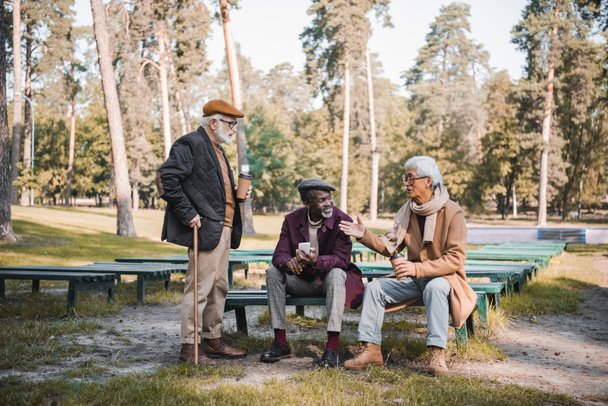 Hombre asiático mayor con café para ir señalando con la mano mientras habla con amigos interracial con teléfono inteligente en el parque  - Foto, Imagen