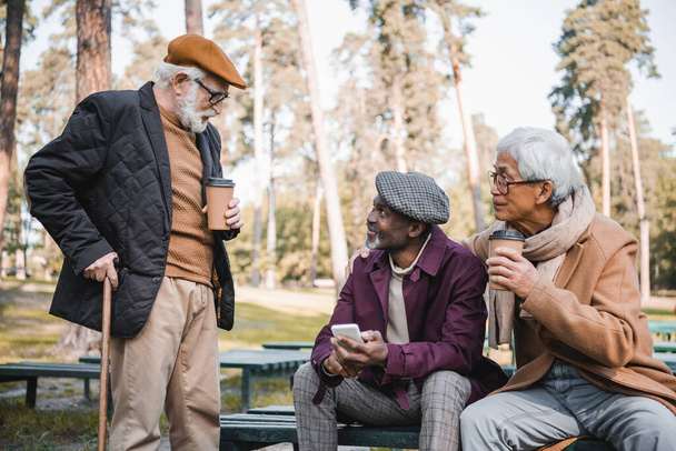 Старший мужчина с тростью, стоящий рядом с межрасовыми друзьями со смартфоном и бумажной чашкой в парке  - Фото, изображение