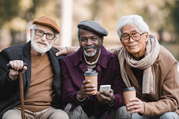 Sorridente anziani amici interrazziale con cellulare e caffè per andare a guardare la fotocamera all'aperto  - Foto, immagini