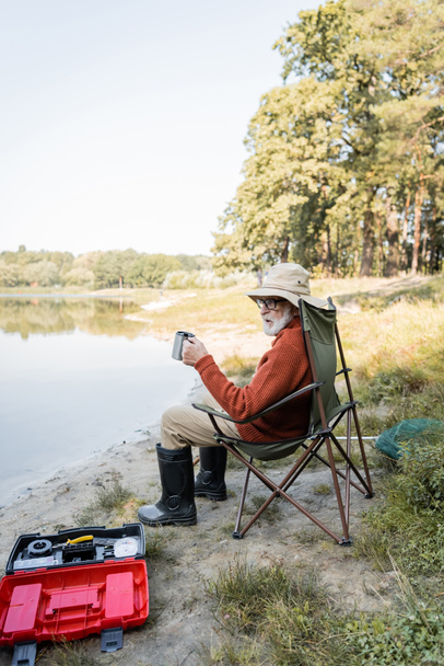 シニア男で釣り服を保持熱カップ近くツールボックスと湖  - 写真・画像